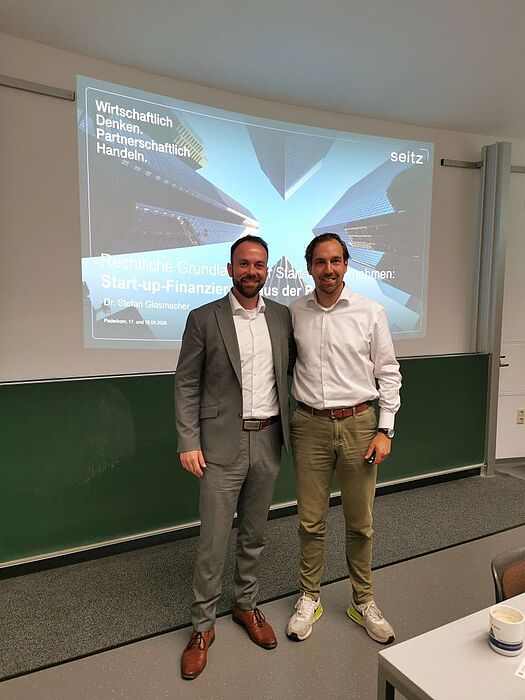 Alternativtext: Prof. Dr. David Bartlitz und Dr. Stefan Glasmacher stehen lächelnd im Hörsaal vor projizierten Veranstaltungsfolien.
