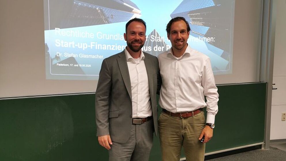 Alternativtext: Prof. Dr. David Bartlitz und Dr. Stefan Glasmacher stehen lächelnd im Hörsaal vor projizierten Veranstaltungsfolien.